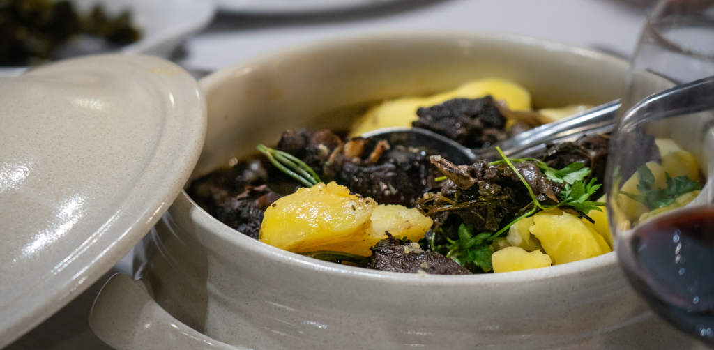 Panela de barro aberta com carne estufada, batatas e ervas, com colher metálica no interior.