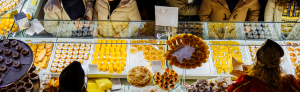 Vitrine com doces amarelos, laranjas e castanhos, com clientes agasalhados do outro lado.