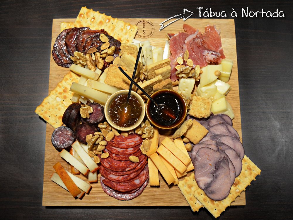 Tábua de madeira com enchidos fatiados, queijos em cubos e palitos, nozes, bolachas e duas taças com mel.