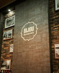 Parede interior com o logótipo “Bijou Restaurante desde 1912”, ladeada por tijolo exposto e fotografias antigas.