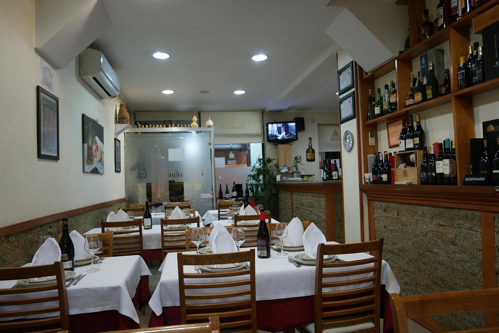 Sala de restaurante com mesas cobertas por toalhas brancas e vermelhas, garrafas de vinho, copos e estante com bebidas ao fundo.