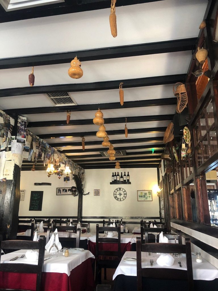 Sala de restaurante com mesas postas, teto com vigas escuras e decoração tradicional com abóboras secas e garrafas.