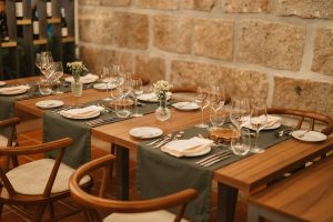 Sala de restaurante com mesas de madeira preparadas com copos de vinho, toalhas verdes e pequenos arranjos florais, junto a parede em pedra.