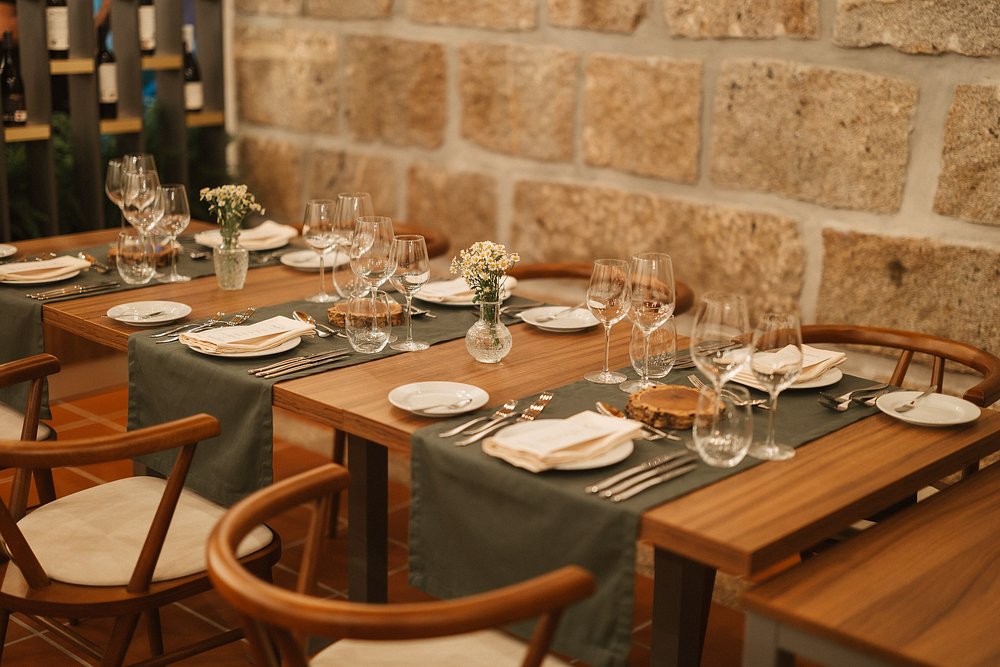 Sala de restaurante com mesas de madeira preparadas com copos de vinho, toalhas verdes e pequenos arranjos florais, junto a parede em pedra.