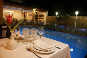 Mesa com toalha branca, copos, pratos e um vaso com tulipa vermelha junto à piscina iluminada.