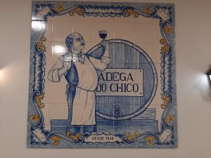 Painel de azulejos azul e branco com a ilustração de uma figura ao lado de um barril com a inscrição “Adega do Chico”, rodeado por moldura decorativa.