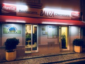 Entrada do Restaurante Baliza com toldo vermelho, portas de vidro e vasos decorativos.
