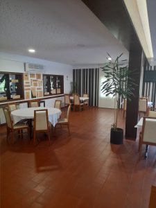 Sala de restaurante com mesas cobertas com toalhas brancas, cadeiras de madeira, expositores de vinho na parede e uma planta decorativa.