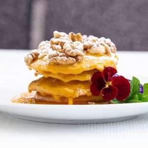 Panquecas empilhadas com nozes por cima, mel, açúcar em pó, flor comestível e hortelã.