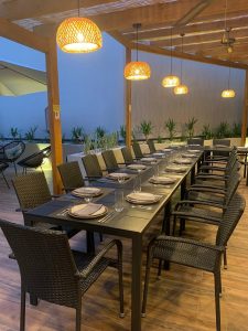 Mesa longa com doze lugares sob uma pérgola de madeira iluminada, com cadeiras escuras e decoração simples.