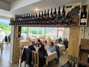 Sala de restaurante com mesas, clientes a comer e garrafeira suspensa com garrafas de vinho.