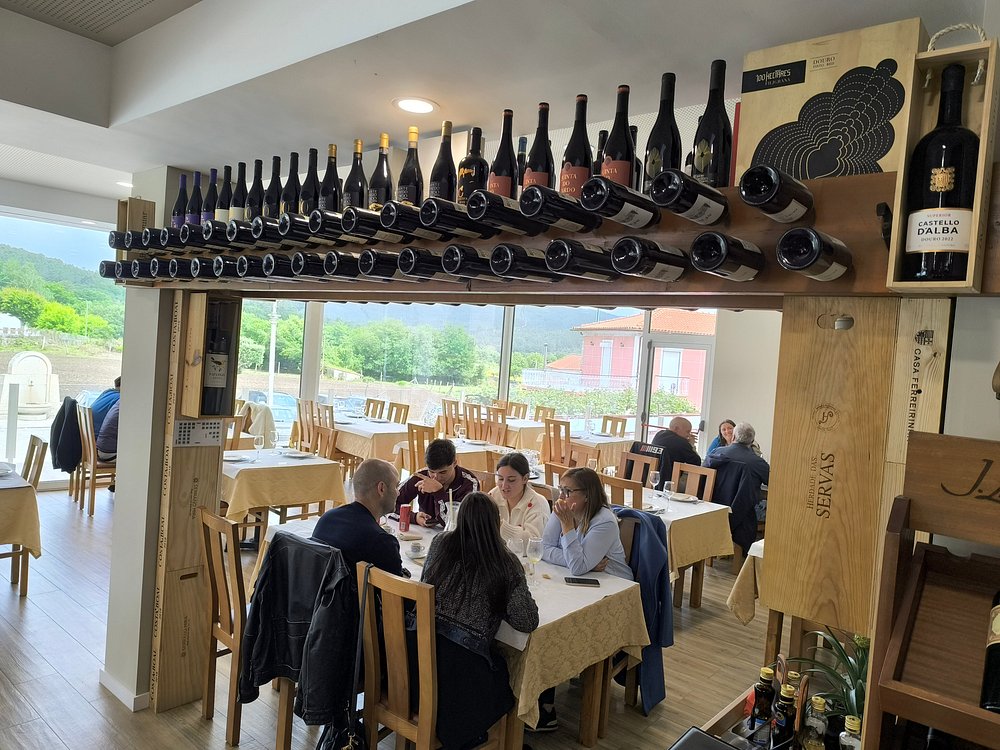 Sala de restaurante com mesas, clientes a comer e garrafeira suspensa com garrafas de vinho.