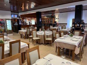 Sala de restaurante com mesas preparadas com toalhas claras, pratos, copos e talheres, com um balcão ao fundo e iluminação suave.