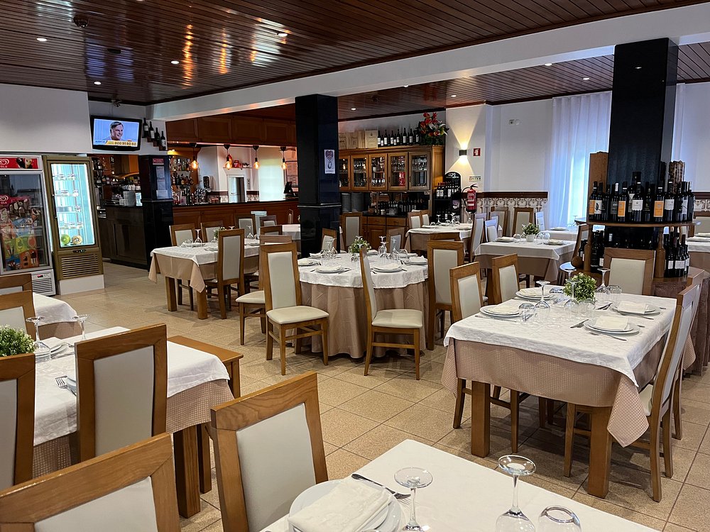 Sala de restaurante com mesas preparadas com toalhas claras, pratos, copos e talheres, com um balcão ao fundo e iluminação suave.