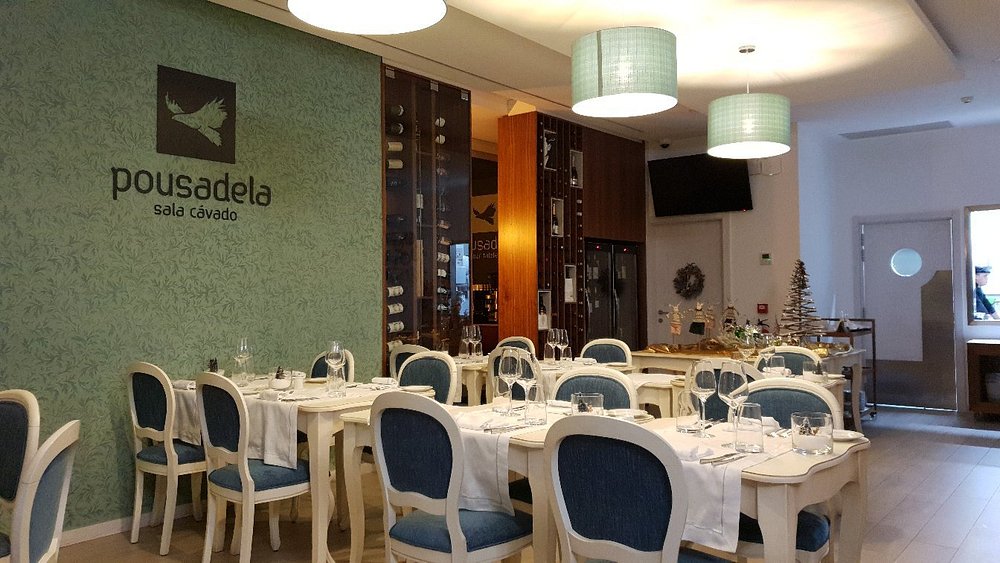 Sala de restaurante com mesas postas, cadeiras estofadas em azul, parede decorativa com o nome Pousadela Sala Cávado e garrafeira ao fundo.
