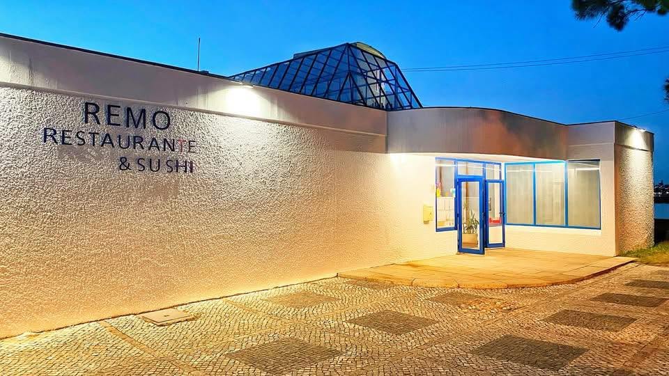 Fachada de um restaurante com parede branca texturizada, janelas e porta azul‑marinho, iluminação exterior e cobertura em vidro em forma de pirâmide ao anoitecer.