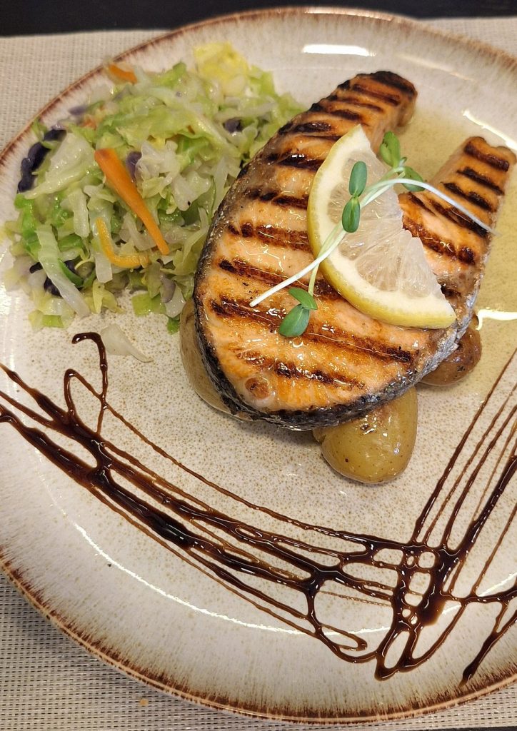 Salmão grelhado com batatas, legumes variados e decoração de molho escuro.