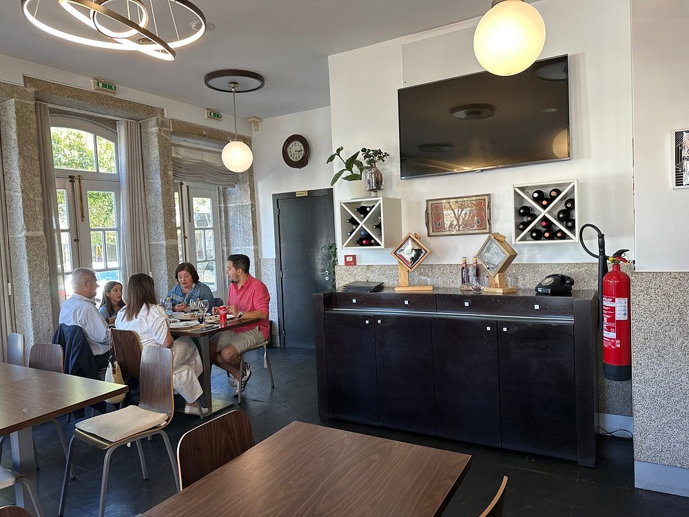 Sala de restaurante com cinco pessoas sentadas à mesa, televisão na parede, garrafeiras, janelas em pedra e decoração moderna.