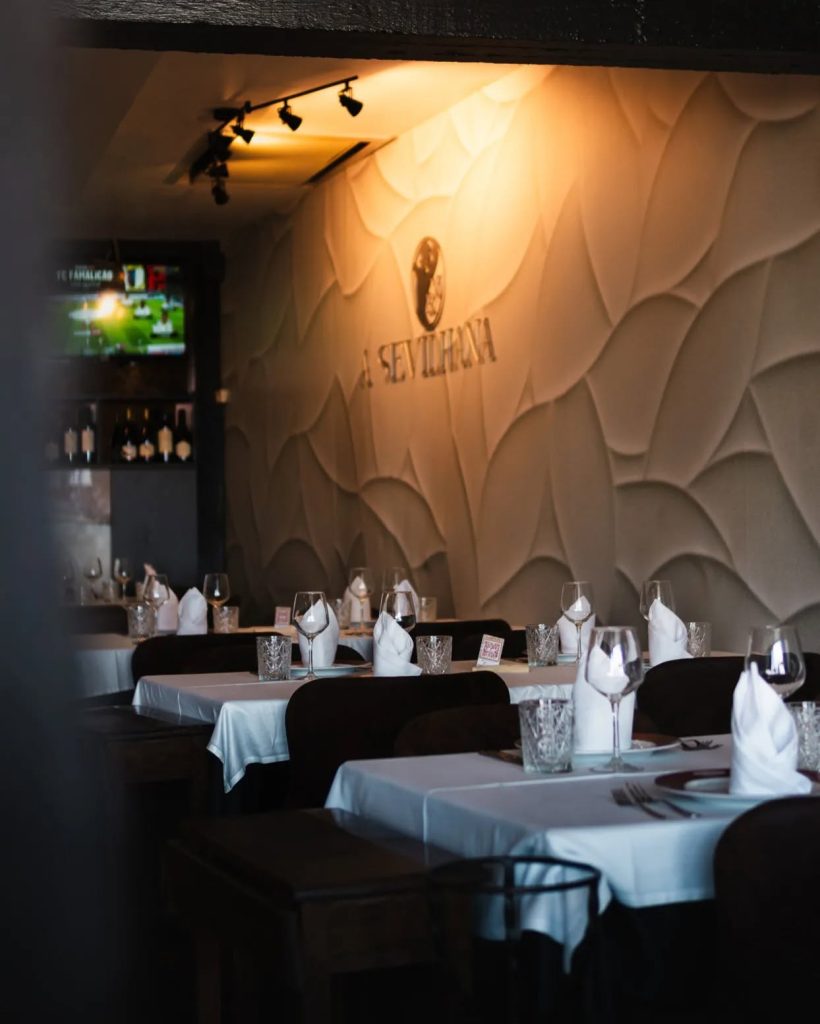 Sala de restaurante com mesas postas, toalhas brancas, copos de vinho e parede decorativa com o nome “A Sevillana”.