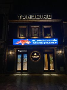 Fachada do Restaurante Tanoeiro à noite, com letreiros iluminados e painel digital com imagem de leitão e contactos.