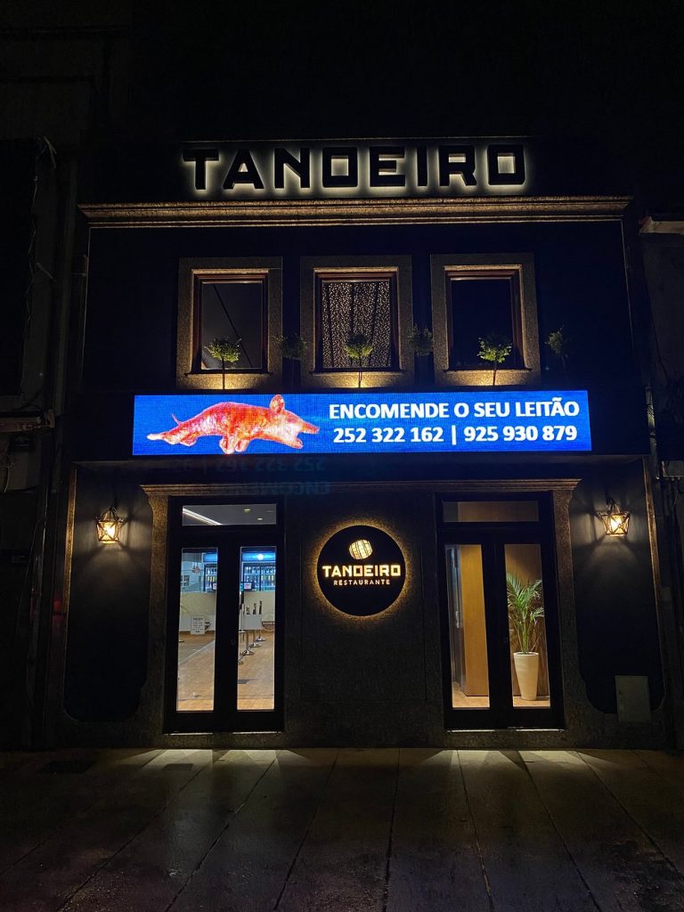 Fachada do Restaurante Tanoeiro à noite, com letreiros iluminados e painel digital com imagem de leitão e contactos.