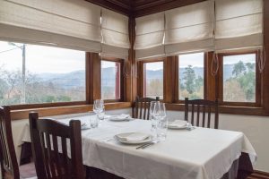 Mesa com toalha branca, quatro lugares completos e janelas grandes com vista para colinas e árvores.
