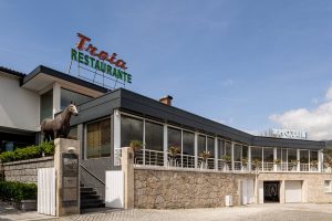 Fachada em pedra e vidro com letreiro “Troia Restaurante” e estátua de cavalo sobre pedestal.