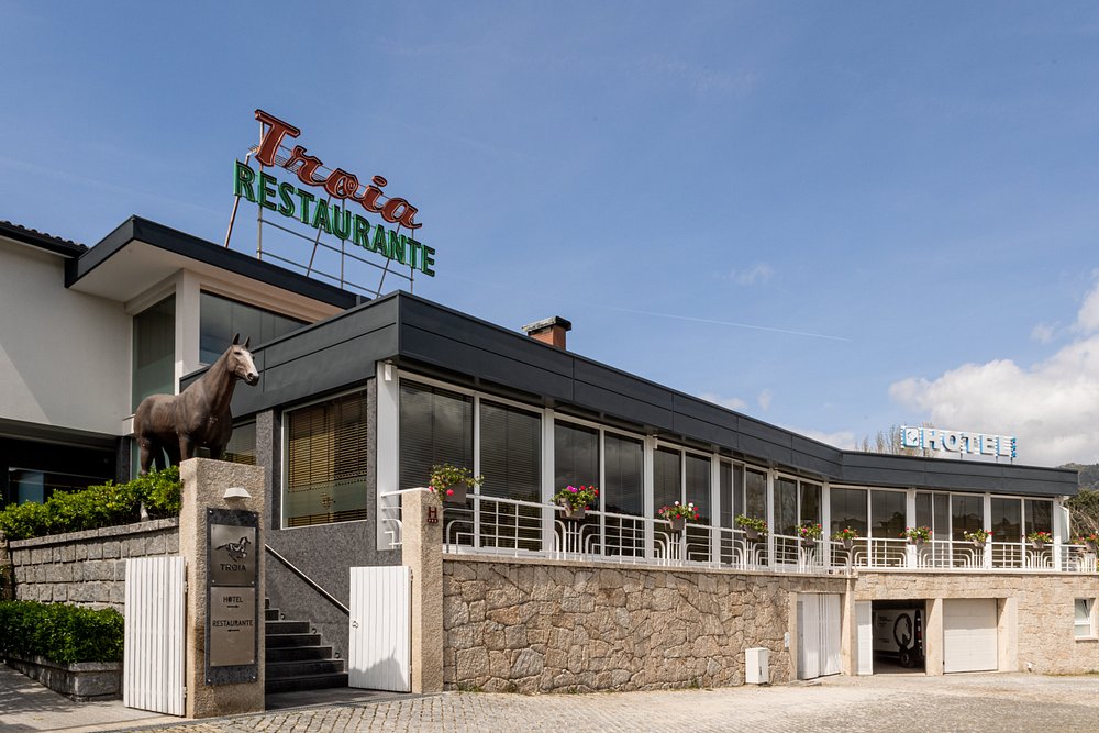Fachada em pedra e vidro com letreiro “Troia Restaurante” e estátua de cavalo sobre pedestal.