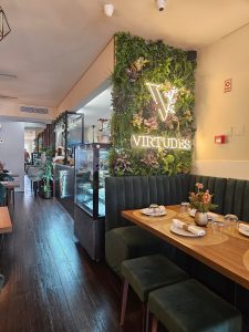 Mesa de restaurante preparada em frente a uma parede verde com plantas e letreiro iluminado “Virtudes”.