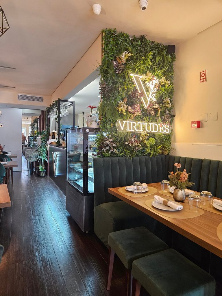 Mesa de restaurante preparada em frente a uma parede verde com plantas e letreiro iluminado “Virtudes”.