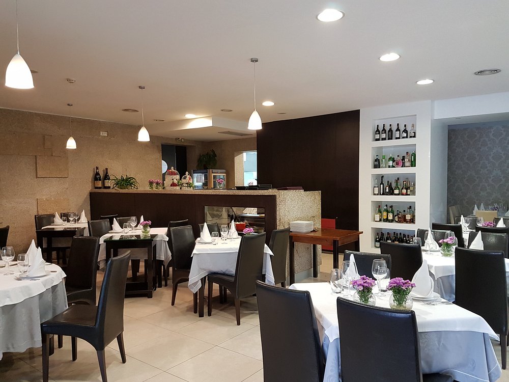 Sala de restaurante com mesas redondas cobertas com toalhas brancas, copos, guardanapos dobrados e pequenas flores roxas.