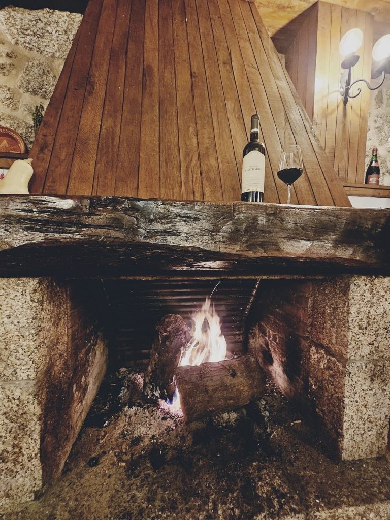 Lareira rústica acesa com garrafa de vinho tinto e copo pousados numa prateleira de madeira.
