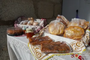 Mesa com pães, bolos, frascos de mel e doces embalados, dispostos sobre toalhas bordadas junto a uma parede de pedra.