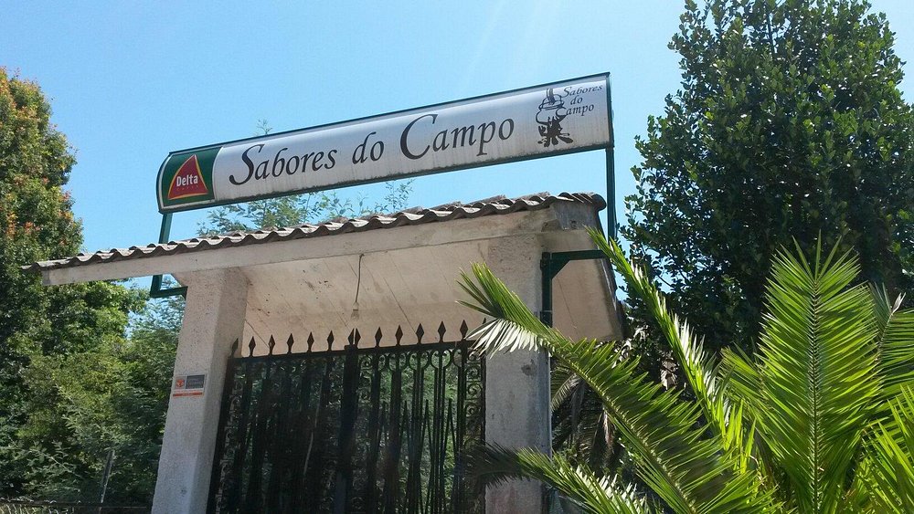 Entrada com portão metálico e letreiro Sabores do Campo sob telhado, rodeada por vegetação.