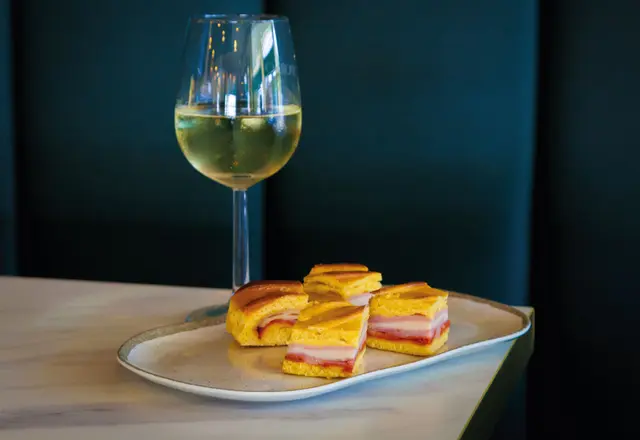 Prato com quatro mini‑sandes retangulares e copo de vinho branco sobre mesa clara.