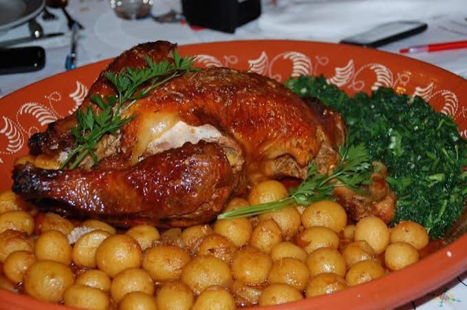 Frango assado inteiro com pele dourada, rodeado de batatas assadas e verduras verdes num prato decorativo.