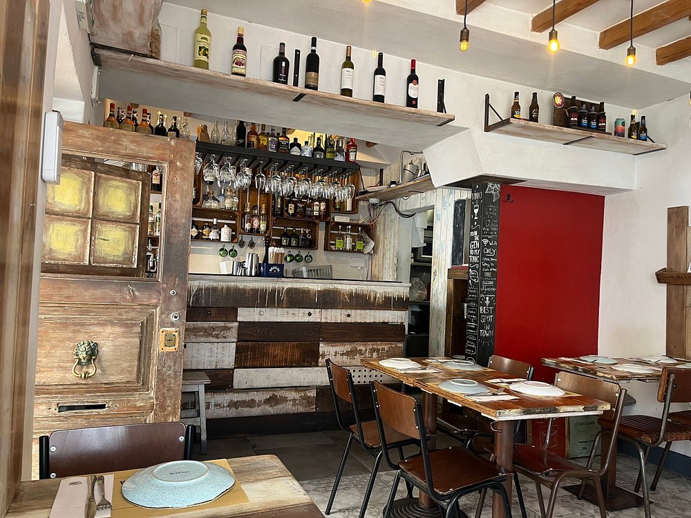Interior de restaurante com balcão em madeira, garrafas expostas, mesas e iluminação quente.