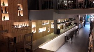 Interior de bar com prateleiras iluminadas com garrafas, balcão em madeira, bancos altos e mezzanine com lugares adicionais.