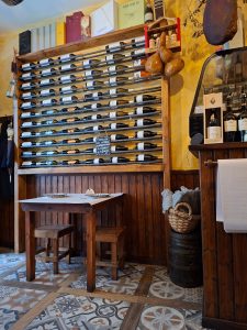Garrafeira em madeira com várias garrafas de vinho e mesa pequena preparada para duas pessoas.