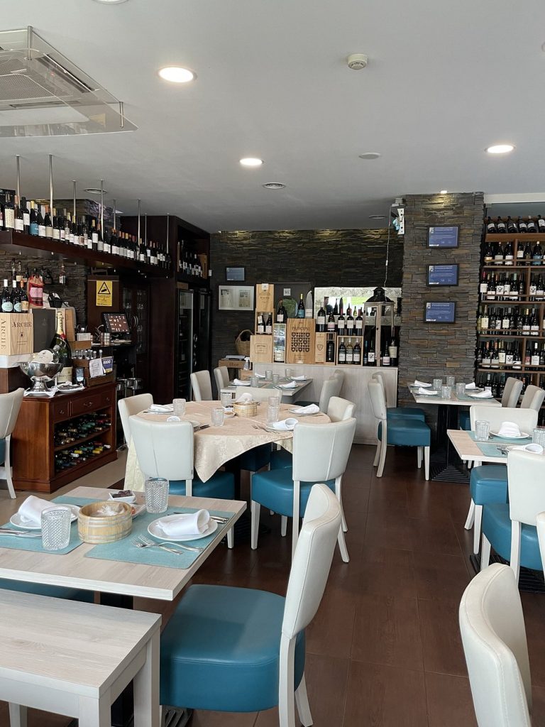 Sala de restaurante com mesas postas, cadeiras brancas e azuis, e garrafeira com várias garrafas de vinho.