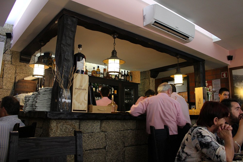 Balcão de restaurante com pessoas ao redor, paredes em pedra, garrafas expostas e iluminação quente.