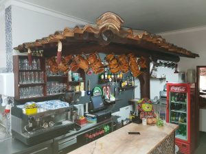 Balcão com máquina de café, copos, garrafas e frigorífico de bebidas, decorado com telhado rústico e canecas penduradas.