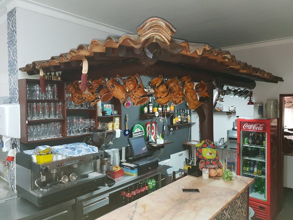 Balcão com máquina de café, copos, garrafas e frigorífico de bebidas, decorado com telhado rústico e canecas penduradas.