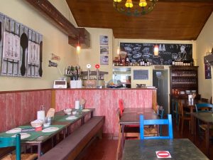 Interior do Tira‑Tapas com mesas de madeira, cadeiras coloridas, bancada com garrafas e decoração alusiva a café, cerveja e tapas.