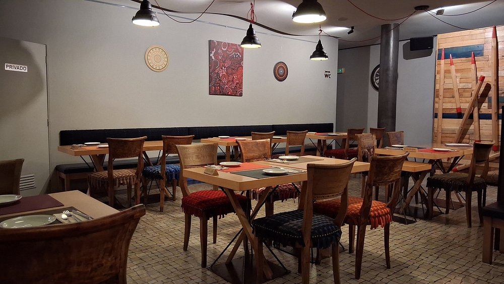 Sala de refeições com mesas de madeira preparadas com pratos, talheres e bases individuais.