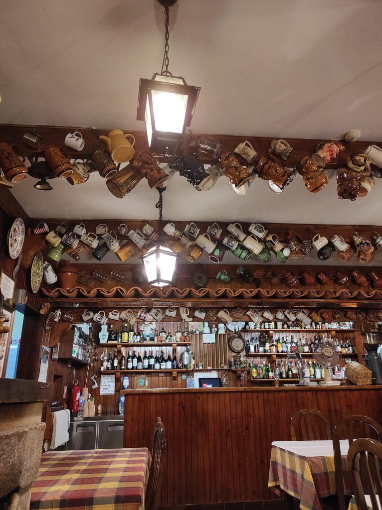 Interior rústico da Adega do Emídio, com balcão em madeira, garrafas expostas e canecas penduradas no teto.