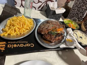 Bife grelhado com alecrim acompanhado por arroz branco, batatas fritas, salada e molho, apresentado numa bandeja do Restaurante Caravela.