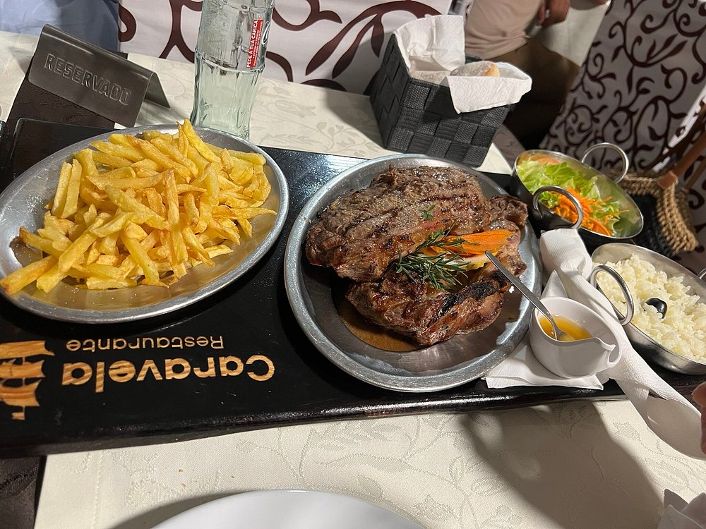Bife grelhado com alecrim acompanhado por arroz branco, batatas fritas, salada e molho, apresentado numa bandeja do Restaurante Caravela.