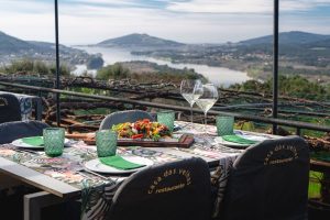 Mesa exterior com toalha estampada, copos verdes, pratos com guardanapos verdes, taças de vinho branco e travessa de salada, com vista para rio e colinas.