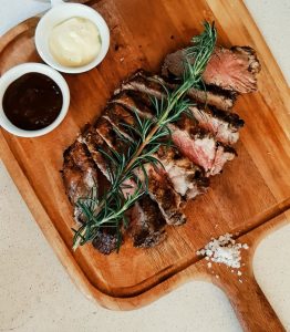 Tábua de madeira com fatias de carne grelhada, ramos de alecrim, dois molhos em taças pequenas e sal grosso.